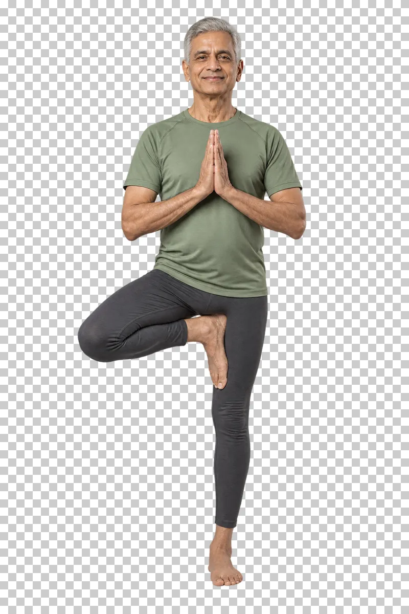 An older man in a sage-green shirt balancing in yoga tree pose with his palms together.
