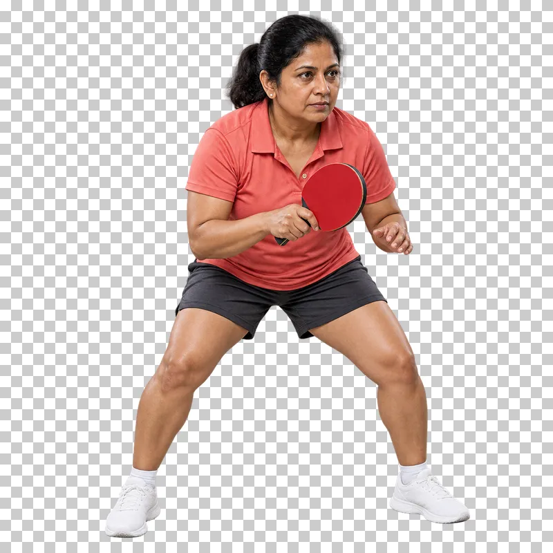 A woman in a coral polo and charcoal shorts holding a table tennis paddle in a low ready stance.