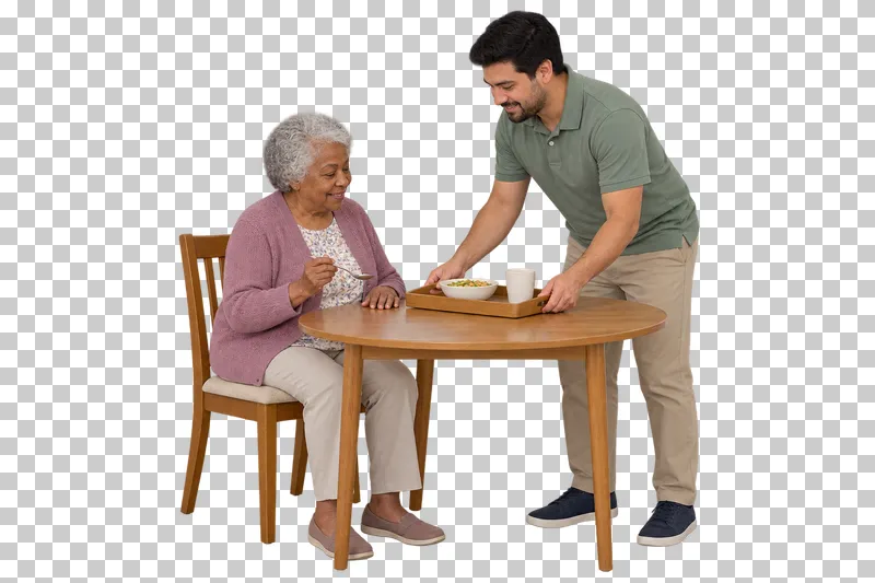 A caregiver sets a meal tray on a table while a seated older woman holds her own spoon.