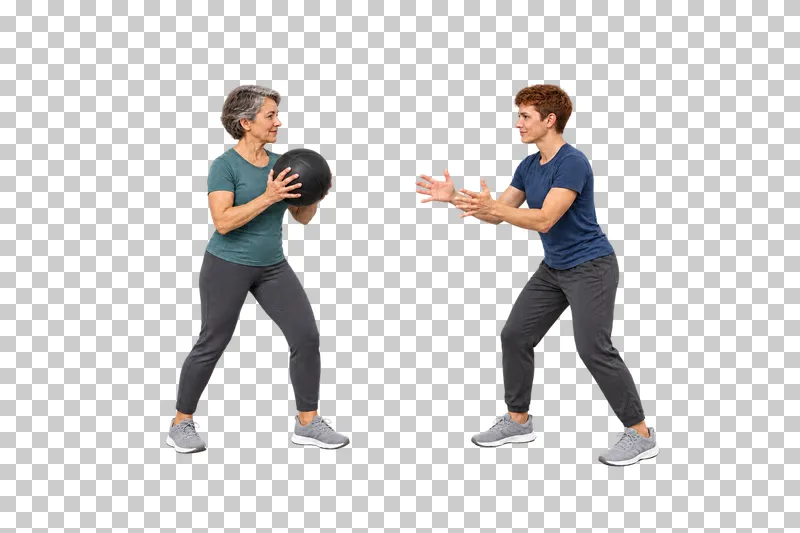 A gray-haired woman holding a medicine ball before passing to a copper-haired adult with open hands.